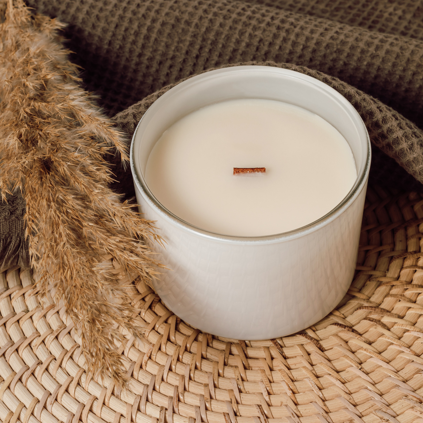 Soy candle handmade with wooden wick in white glass jar with brown towel and pampas grass at the background